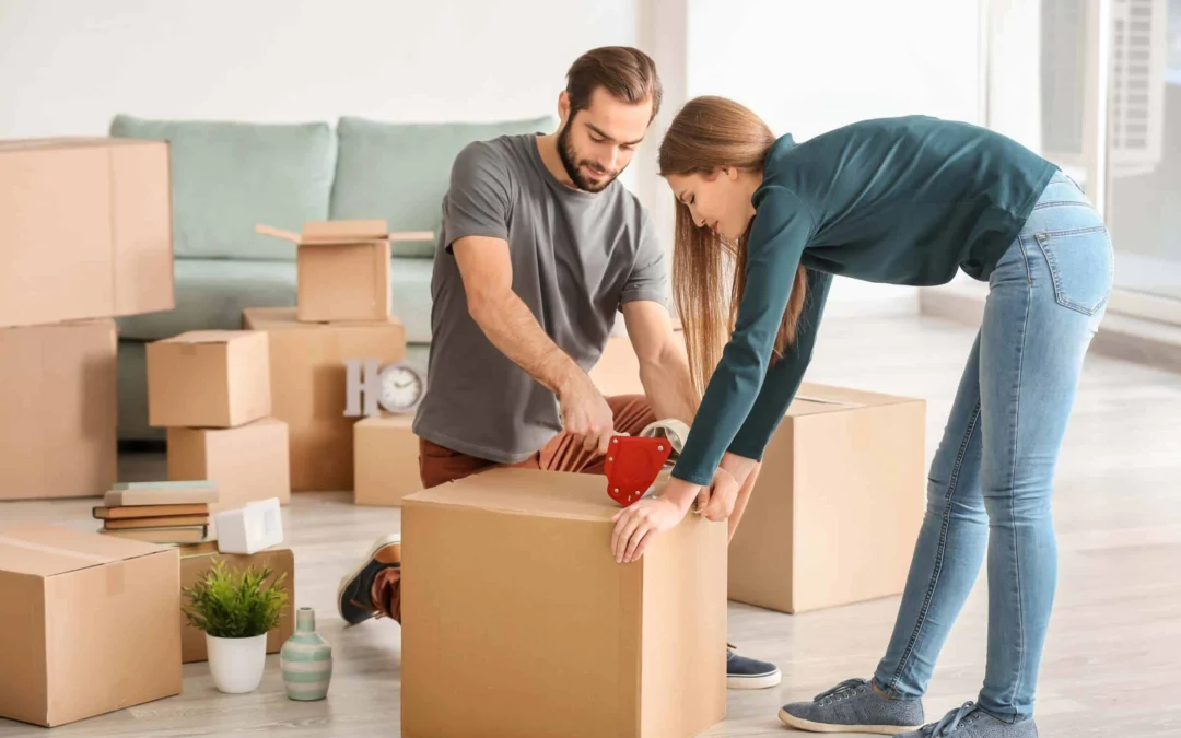 Couple sealing a cardboard box with tape during a home move in a bright living room.