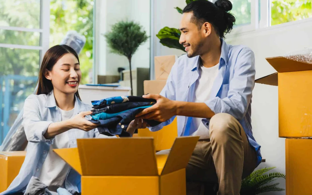 Two friends packing folded clothes into a cardboard box in a sunlit room with moving boxes nearby.