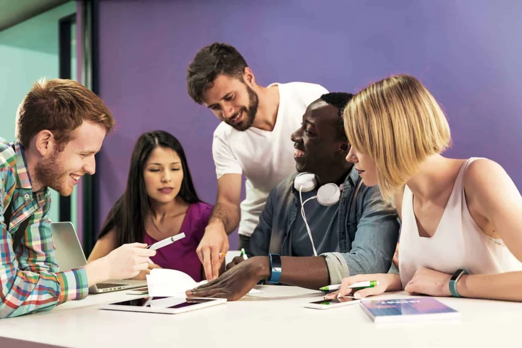 Five young adults of different backgrounds collaborate around a white table, examining papers and devices together.