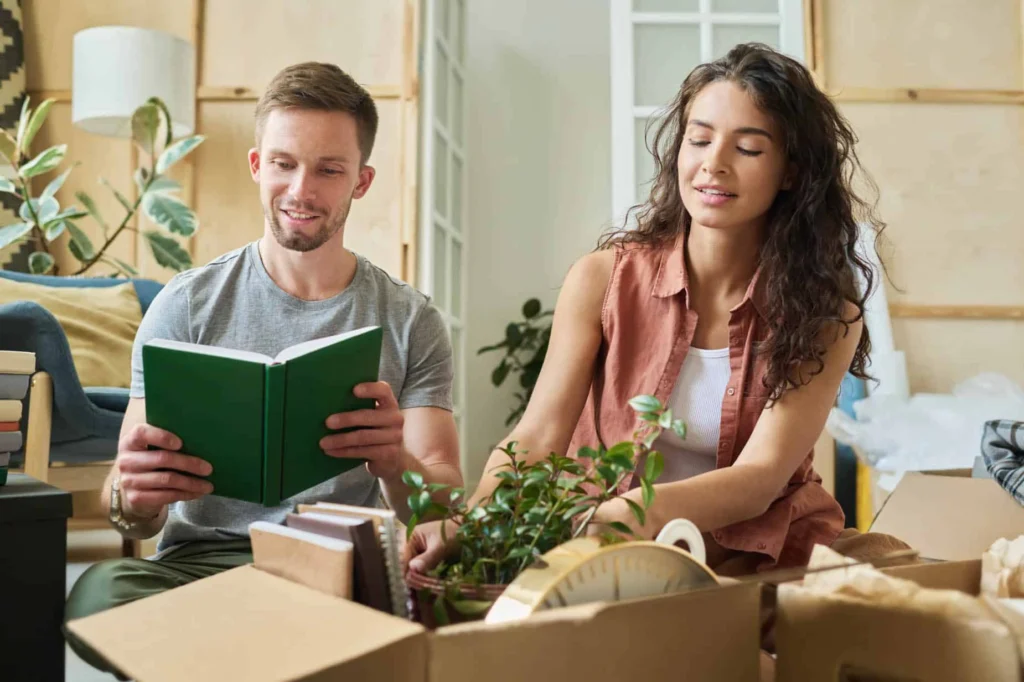 Couple sits among moving boxes, one reads a green notebook while the other sorts plants for their move home.