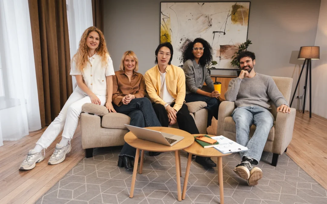 Five adults sit on a beige sofa in a modern living room, smiling at the camera with a laptop and magazines on the coffee table.